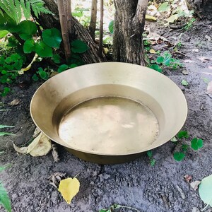 May include: A large, round, gold-colored metal bowl sits on a bed of dirt and leaves. The bowl is empty and has a smooth, reflective surface.