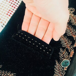 Op de afbeelding: Zwart fluwelen jasje met gouden borduurwerk van bladeren en een knoop. Het jasje heeft een zak aan de linkerkant.