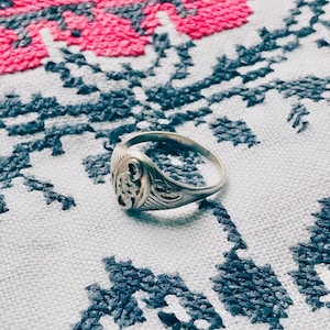 May include: A silver ring with an ornate design on the top. The ring is sitting on a white fabric with a pink and blue floral pattern.
