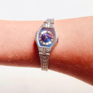 May include: A silver wristwatch with a blue face and a silver band. The watch has a unique octagonal shape and a decorative design on the band.