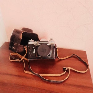 May include: Vintage black and silver camera with a leather case and strap. The camera has a lens cap and is sitting on a wooden surface. The case is open, revealing the camera's body. The strap is tan and black.