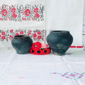 Può includere: Due vasi di terracotta neri con una ghirlanda di papaveri rossi davanti a loro. I vasi sono appoggiati su una tovaglia bianca con disegni floreali ricamati.