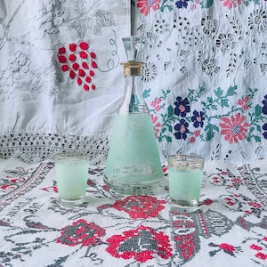May include: A clear glass bottle with a green liquid and a silver stopper, sitting on a white tablecloth with red and gray floral embroidery. Two small shot glasses filled with the same green liquid are on either side of the bottle.