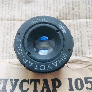 K&ouml;nnte beinhalten: Ein schwarzes Kameraobjektiv mit dem Text "INDUSTAR 105" und "4/50" auf der Seite. Das Objektiv hat ein blaues Glaselement, das durch die &Ouml;ffnung sichtbar ist.