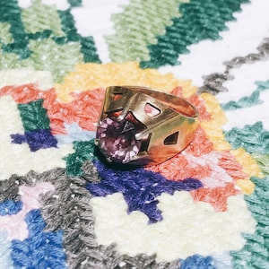 May include: A gold ring with a large, faceted purple gemstone set in the center. The ring has a geometric design with intricate details. The ring is resting on a colorful, patterned fabric.