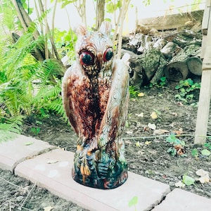 May include: A ceramic owl statue with brown, green, and white paint. The owl has large, red eyes and intricate details on its feathers. The statue is standing on a base with a floral design.