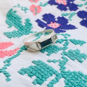 May include: A silver ring with a dark blue rectangular stone and a small clear stone on the side.