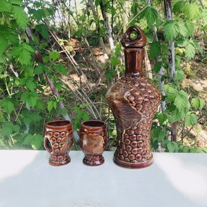 May include: A set of three brown ceramic bottles with a grape design. The bottles have a textured surface and a rounded shape. The largest bottle has a lid with a handle.