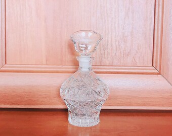 May include: A clear glass bottle with a decorative, faceted design. The bottle has a round body and a short, wide neck.