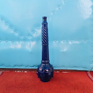 May include: A dark blue glass bottle with a decorative design and a round, studded base. The bottle is standing on a red rug.