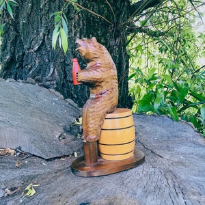 May include: A hand-carved wooden bear figurine holding a red bottle, seated on a light brown barrel. The bear is brown with visible wood grain, and the barrel has black stripes. The figurine sits on a dark brown base, showcasing a rustic, handcrafted aesthetic.