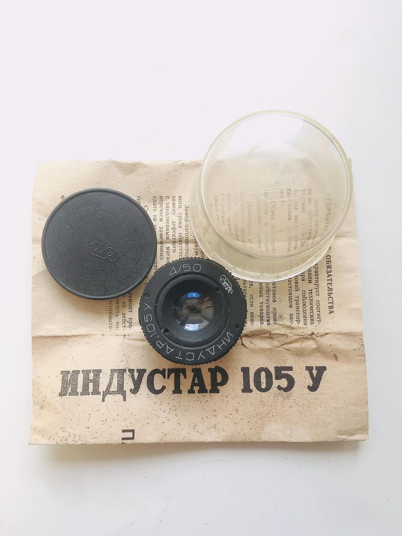 K&ouml;nnte beinhalten: Ein schwarzes Kameraobjektiv mit dem aufgedruckten Text "INDUSTAR 105 Y". Das Objektiv ist von wei&szlig;em Papier mit russischem Text und einem durchsichtigen Plastikdeckel umgeben.