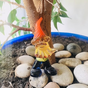 Puede incluir: Una pequeña figura de gnomo de vidrio con un sombrero naranja, cuerpo amarillo, piernas azules y zapatos negros. El gnomo está de pie sobre una cama de pequeñas piedras lisas.