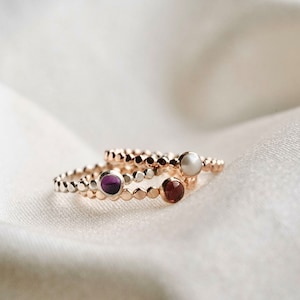 May include: Three delicate rings with beaded bands are displayed. Each ring features a different gemstone: a deep purple, a rich red, and a white pearl. The rings are set against a soft, white background.