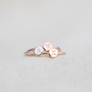 May include: Three delicate rings in silver, rose gold, and gold. Each ring features a small, round charm with an engraved design: the letter "R", a heart, and the letter "K". The rings are stacked together on a soft, white surface.