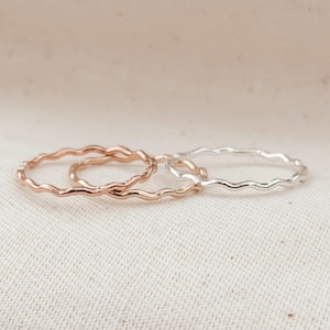 May include: Three thin, delicate rings with a wavy, undulating design. Two of the rings are finished in a warm rose gold tone, while the third ring is finished in a bright, polished silver tone. Each ring features a continuous, smooth texture.