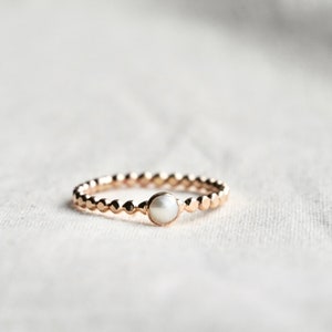 May include: A gold ring with a central white pearl. The band is decorated with small, evenly spaced gold beads. The ring is set against a soft, neutral background, highlighting its elegant design.