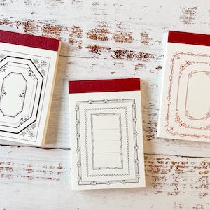 May include: Three decorative notepads with claret tops and ornate border designs. The notepads feature different frame styles in black and red ink on a cream background. The notepads are arranged on a distressed white wooden surface.