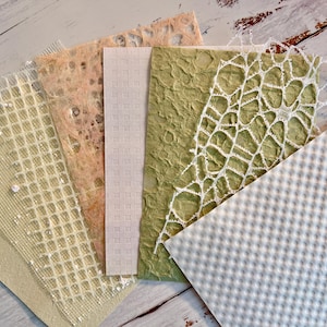 May include: A collection of handmade paper samples in various textures and colors. The samples include a light green paper with a textured surface, a white paper with a grid pattern, and a peach-colored paper with a delicate, open weave.