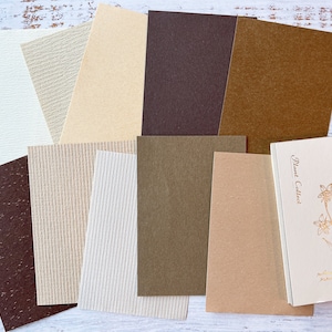 May include: A collection of 11 different textured paper sheets in various shades of brown, beige, and white. The sheets are arranged in a staggered pattern on a white wooden surface.