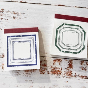 May include: Two small white notebooks with red borders. The notebook on the left has a blue and white patterned border with a blank space inside. The notebook on the right has a green and white patterned border with a blank space inside.