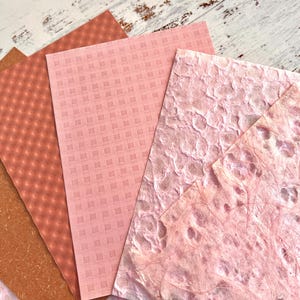 May include: A collection of textured paper sheets in shades of pink and brown. The papers feature various patterns, including a grid, a diamond pattern, and a textured, organic design. The papers are arranged on a light-colored wooden surface.