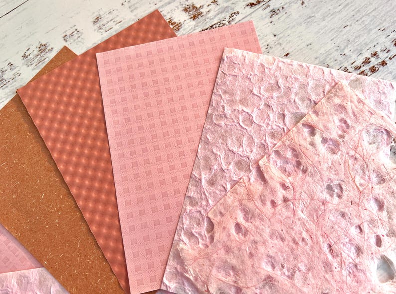 May include: A collection of textured paper sheets in shades of pink and brown. The papers feature various patterns, including a grid, a diamond pattern, and a textured, organic design. The papers are arranged on a light-colored wooden surface.