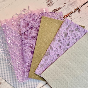 May include: A collection of handmade paper sheets in various textures and colors. The assortment includes sheets in shades of lavender, light gray, and tan. Some sheets feature intricate patterns, including lace-like designs and speckled textures.