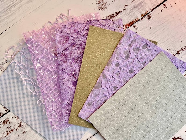 May include: A collection of handmade paper sheets in various textures and colors. The assortment includes sheets in shades of lavender, light gray, and tan. Some sheets feature intricate patterns, including lace-like designs and speckled textures.