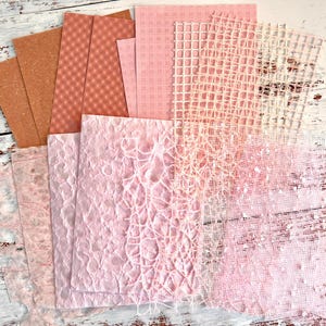 May include: A collection of various textured papers in shades of pink, peach, and brown. The papers include patterned, woven, and textured surfaces, ideal for crafts and scrapbooking. The papers are arranged on a distressed white wooden surface.