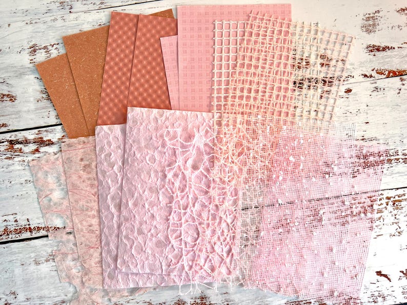 May include: A collection of various textured papers in shades of pink, peach, and brown. The papers include patterned, woven, and textured surfaces, ideal for crafts and scrapbooking. The papers are arranged on a distressed white wooden surface.
