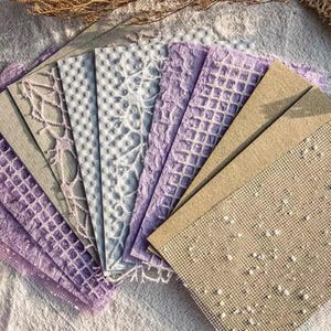 May include: A collection of textured paper sheets in various shades of purple, gray, and beige. The papers feature embossed patterns like lattice, dots, and floral designs, arranged on a white surface with lace accents.