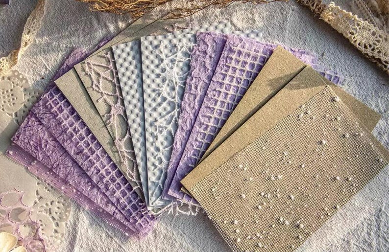 May include: A collection of textured paper sheets in various shades of purple, gray, and beige. The papers feature embossed patterns like lattice, dots, and floral designs, arranged on a white surface with lace accents.