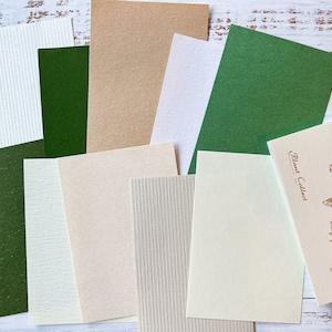 May include: A collection of 10 different textured paper sheets in various colours, including green, beige, and white. The sheets are arranged in a random pattern on a white wooden surface.