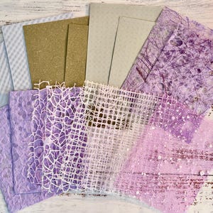 May include: A collection of various textured papers in shades of white, tan, and purple. The papers include patterned, woven, and speckled designs, suitable for crafts and scrapbooking. The arrangement is fanned out, showcasing the different textures and colors.