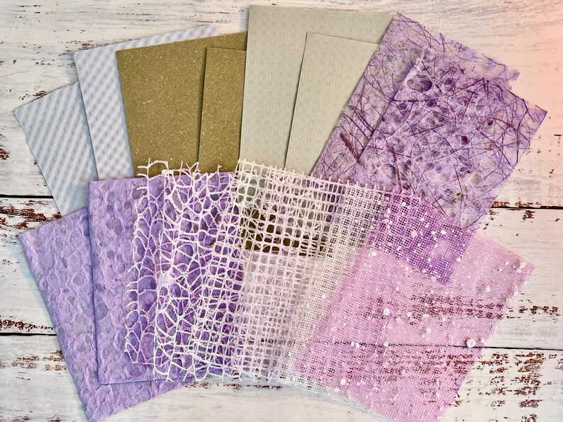 May include: A collection of various textured papers in shades of white, tan, and purple. The papers include patterned, woven, and speckled designs, suitable for crafts and scrapbooking. The arrangement is fanned out, showcasing the different textures and colors.
