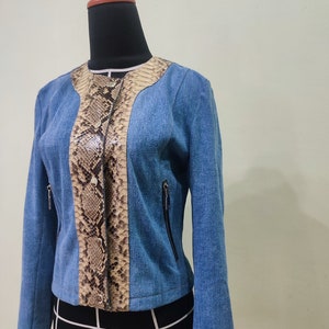 Op de afbeelding: Een blauwe denimjas met een slangenhuidpatroon op de voorkant. De jas heeft een ritssluiting en twee zakken aan de voorkant.