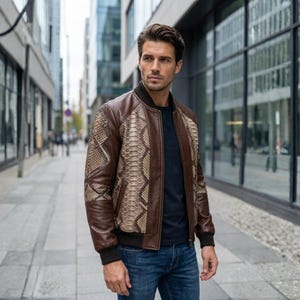 May include: A brown leather bomber jacket with snake skin pattern on the shoulders and upper sleeves. The jacket has a black collar and cuffs, and is worn over a navy blue shirt and blue jeans.