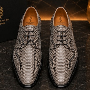 May include: A pair of handmade lace-up shoes crafted from genuine python skin in a grey and black scale pattern. The shoes feature black laces, a tan leather interior lining, and a black sole. The insole is stamped with a snake logo and text.
