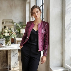 Pode incluir: Uma mulher usando uma jaqueta magenta com estampa de pele de cobra, uma blusa preta e calças pretas. A jaqueta tem fecho de zíper e design sem gola. O look é complementado com joias douradas.