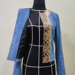 Op de afbeelding: Een blauwe denimjas met een slangenvellenpaneel aan de voorkant. De jas heeft een ritssluiting en knoopaccenten.