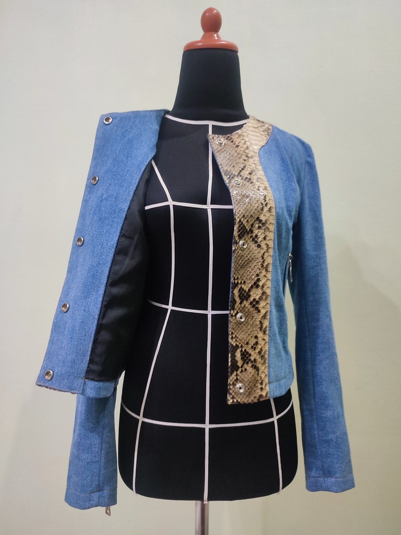 Op de afbeelding: Een blauwe denimjas met een slangenvellenpaneel aan de voorkant. De jas heeft een ritssluiting en knoopaccenten.