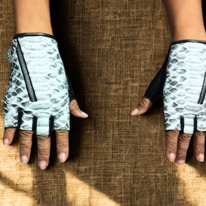 Könnte beinhalten: Ein Paar weiße fingerlose Handschuhe mit einem schwarzen Schlangenhautmuster und einem Reißverschlussdetail am Handgelenk.