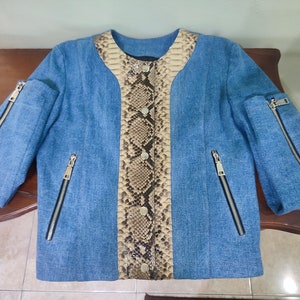 Op de afbeelding: Een blauwe denimjas met een slangenvellenpaneel aan de voorkant. De jas heeft ritszakken aan de zijkanten en mouwen.