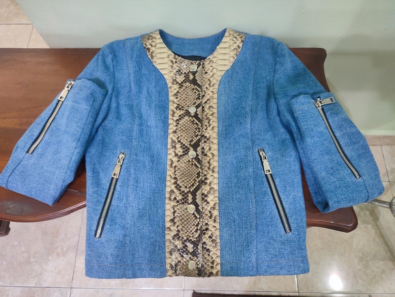 Op de afbeelding: Een blauwe denimjas met een slangenvellenpaneel aan de voorkant. De jas heeft ritszakken aan de zijkanten en mouwen.