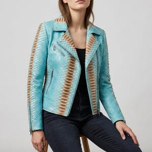 Pode incluir: Casaco de couro turquesa com padrão de pele de cobra e detalhes castanhos nas mangas. O casaco tem um estilo motard clássico com gola de lapela, detalhes de zíper e um corte sob medida. A pessoa está vestindo um top preto e jeans azul escuro.