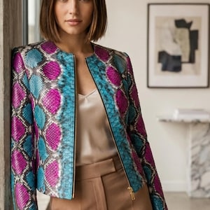Pode incluir: Casaco com zíper e padrão colorido. O padrão apresenta tons de azul, rosa e roxo. O casaco é usado sobre uma parte de cima e calças de cores neutras. O padrão do casaco lembra pele de cobra.