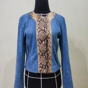 Op de afbeelding: Een blauw denim jack met een slangenhuidpatroon op de voor- en achterkant. Het jack heeft een ritssluiting en een knoopsluiting aan de voorkant.