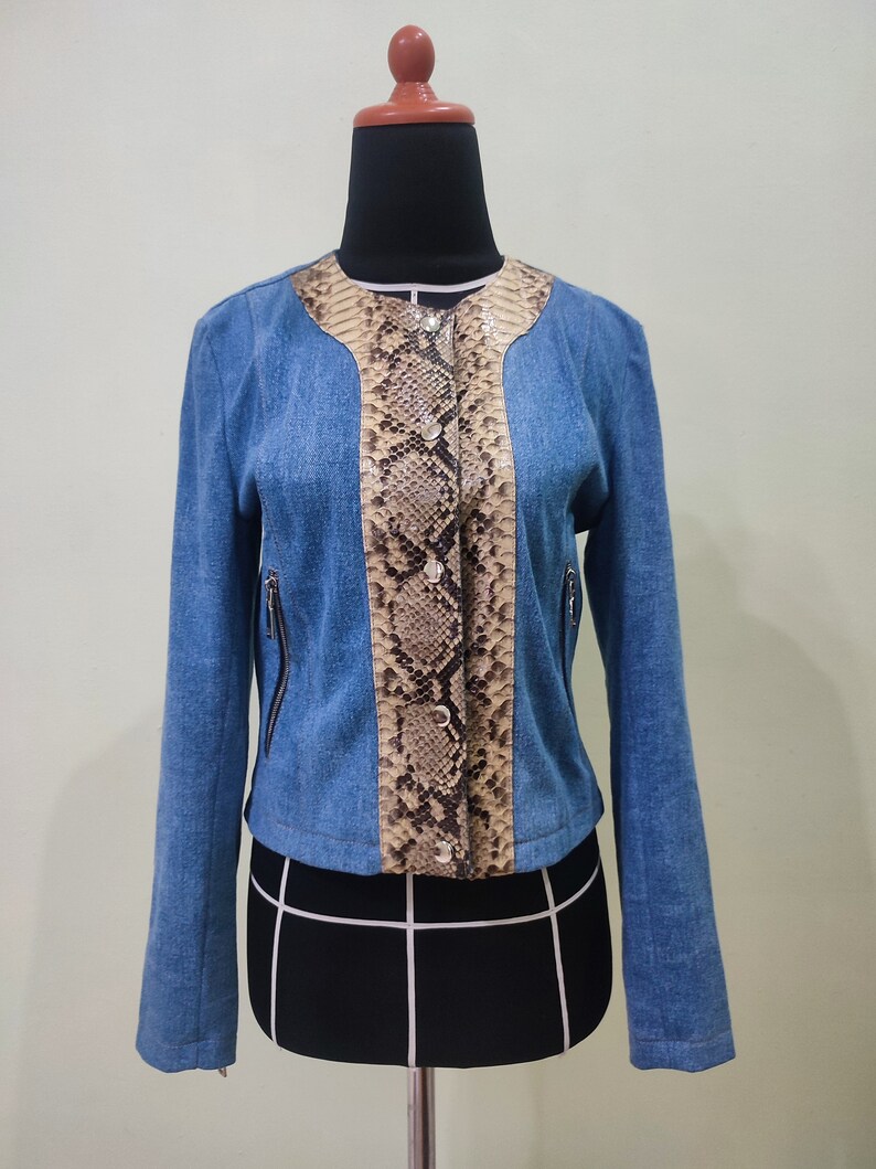 Op de afbeelding: Een blauw denim jack met een slangenhuidpatroon op de voor- en achterkant. Het jack heeft een ritssluiting en een knoopsluiting aan de voorkant.