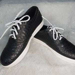 Peut inclure: Une paire de baskets noires avec un motif texturé en forme d'écailles. Les chaussures ont des lacets et des semelles blancs. Les baskets sont présentées sur une surface blanche et moelleuse.